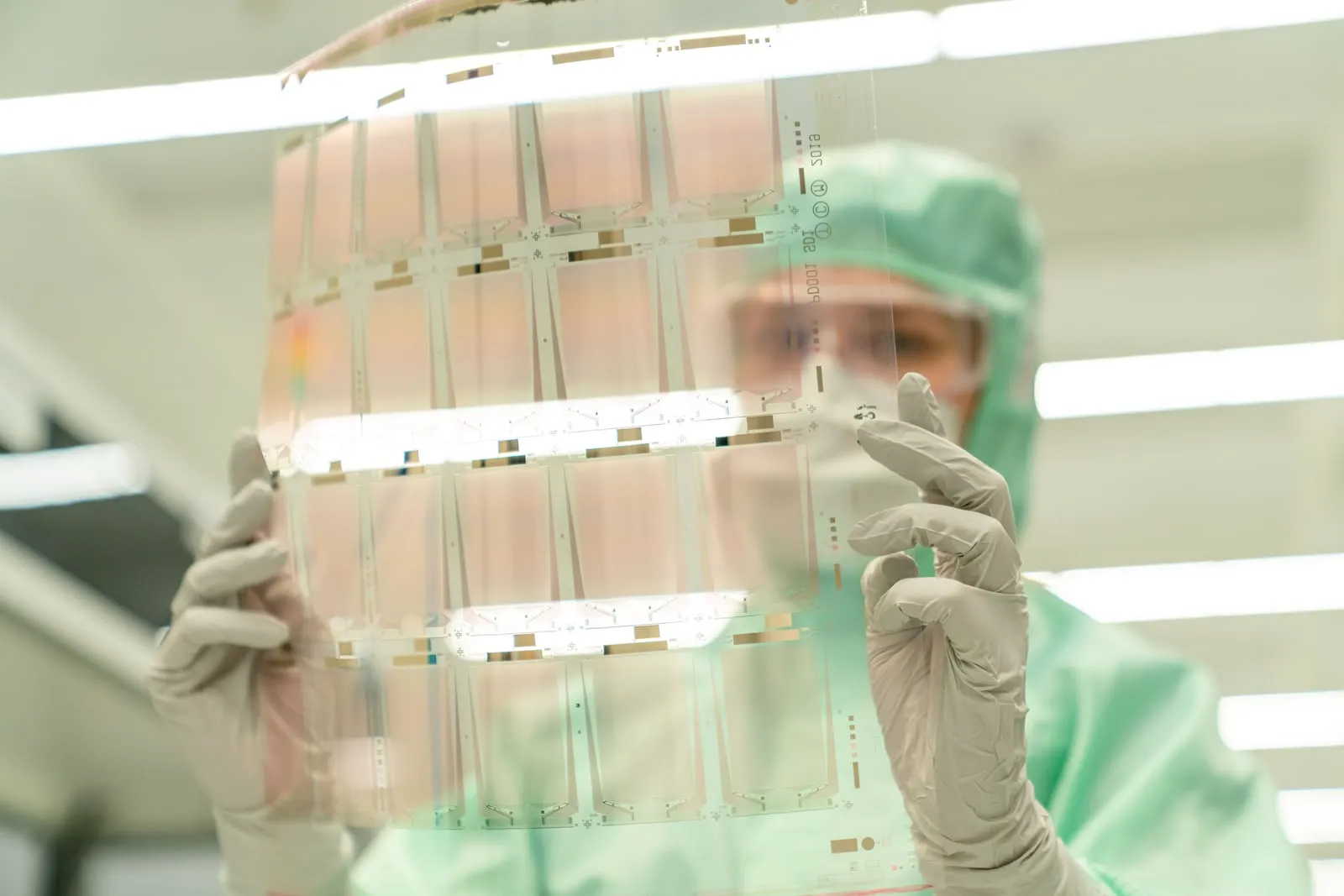 A worker in cleanroom clothing holding a transparent substrate panel up to the light