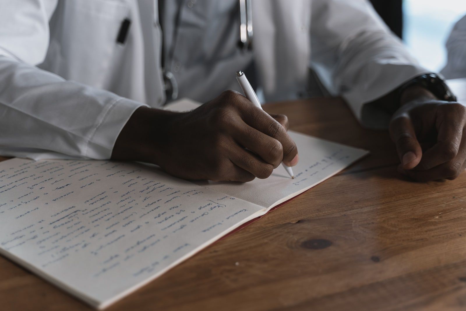 A clinician in a white coat writing in a notebook at a desk