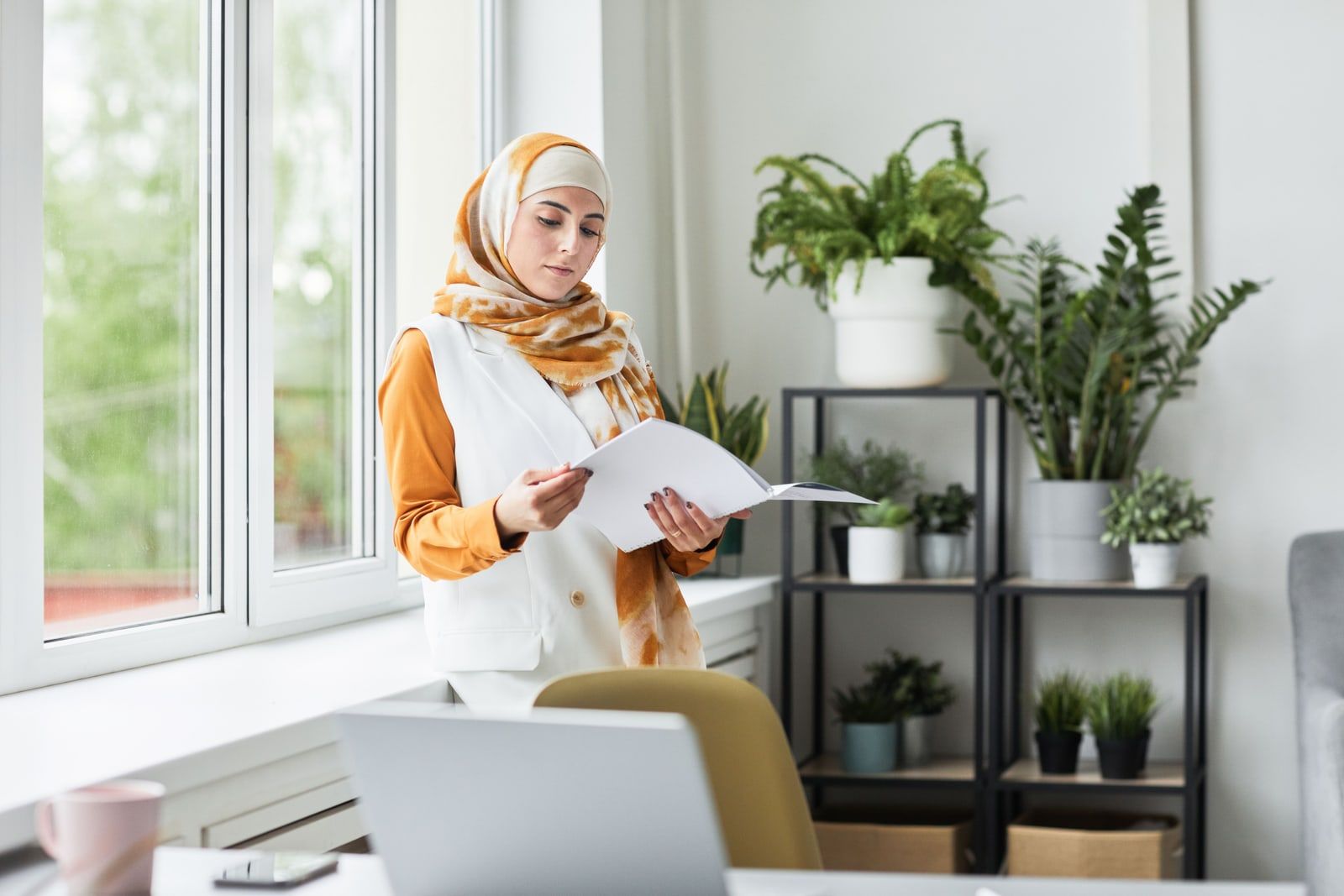 A woman standing by a window reading a printed document in a bright office
