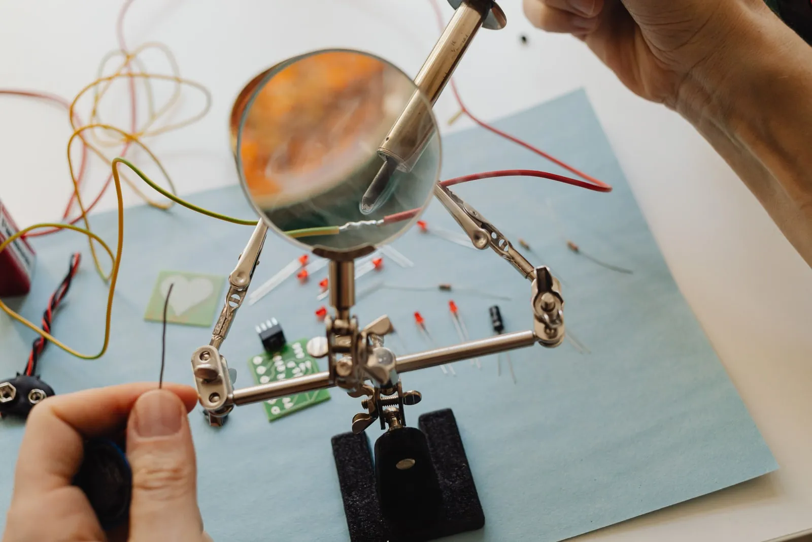 Hands soldering a wire held in a magnifier stand, with components laid out alongside