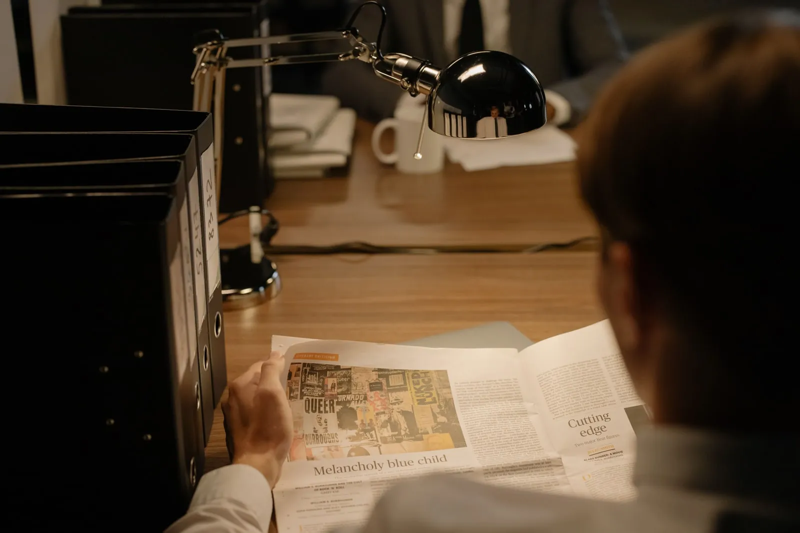 A person reading a newspaper at a desk under a lamp