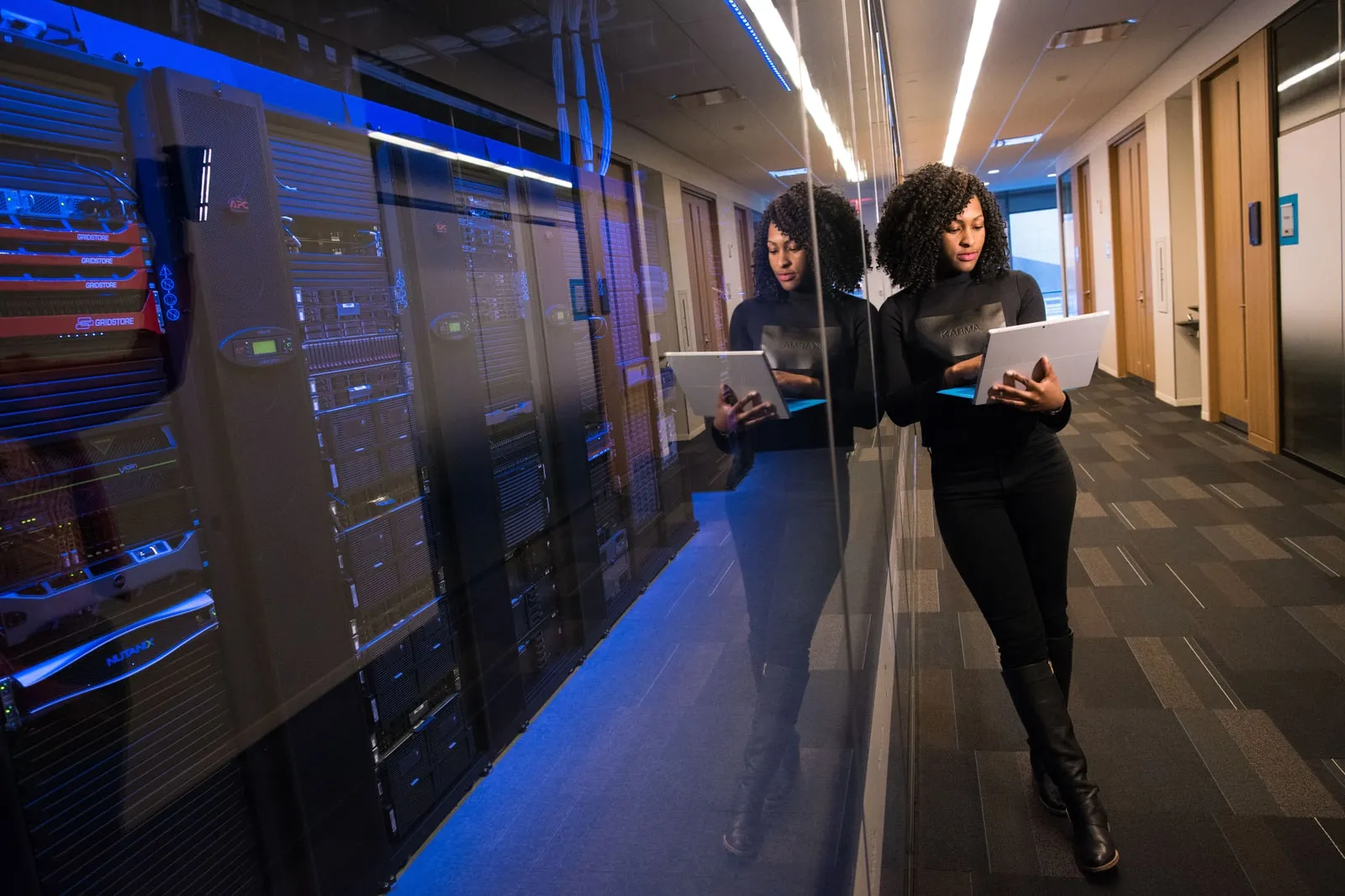 A person with a laptop standing beside a glass-walled server room