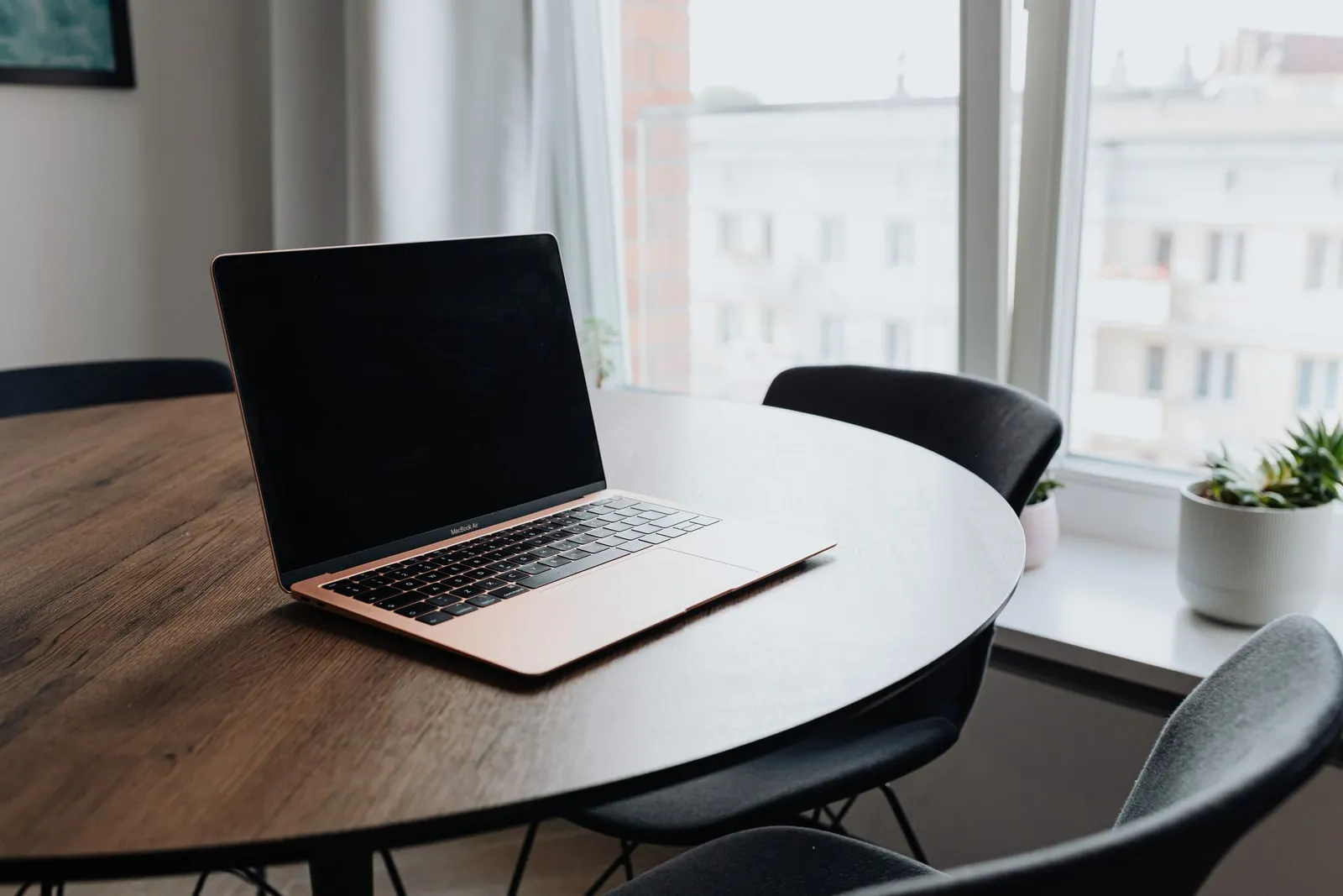 A laptop with a dark screen on a round wooden table by a window