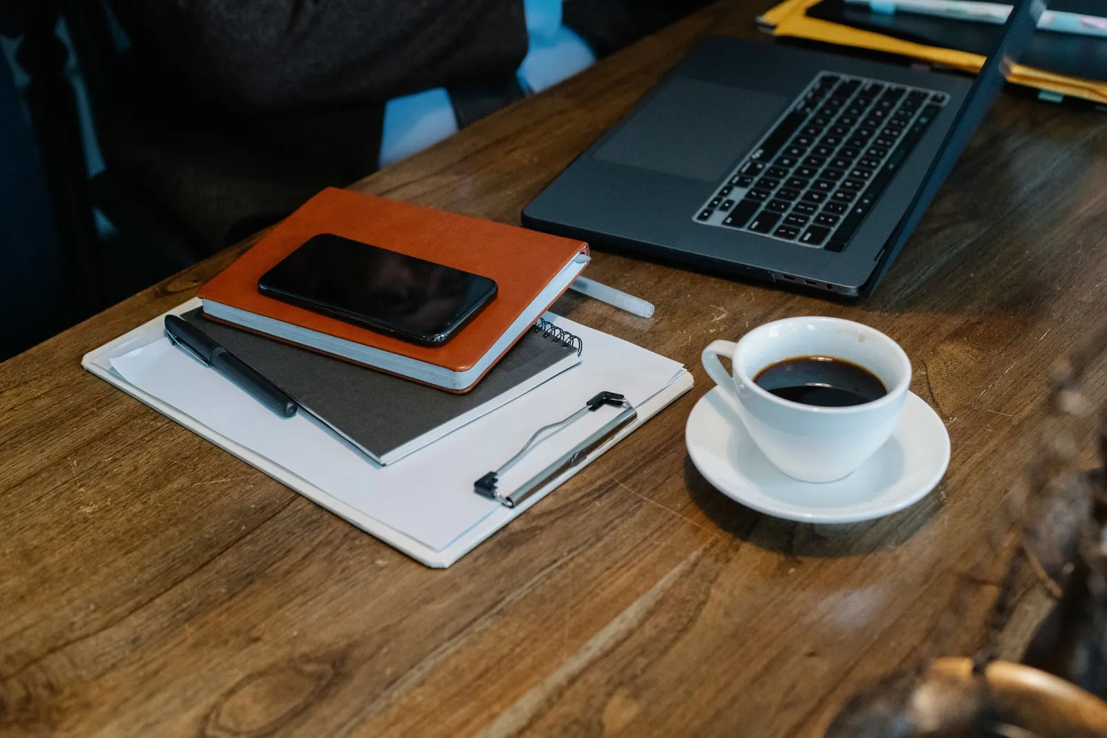 A phone resting on a notebook beside a laptop and a cup of coffee on a wooden table