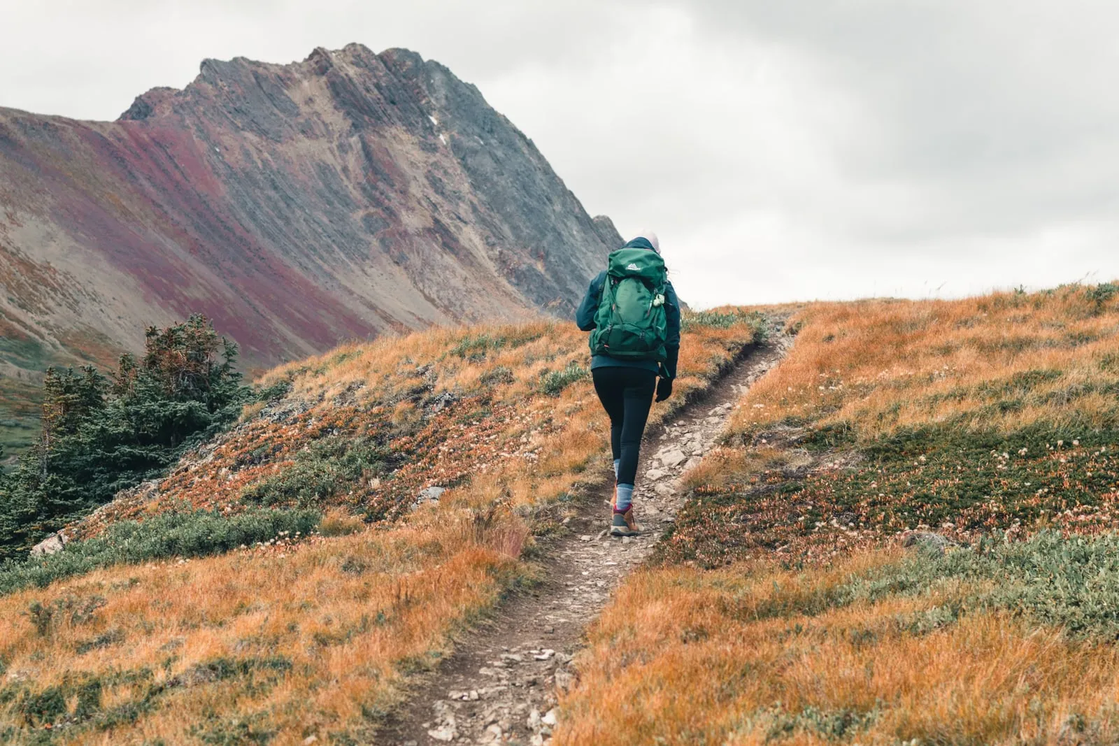 A hiker with a backpack walking a mountain trail below a rocky ridge