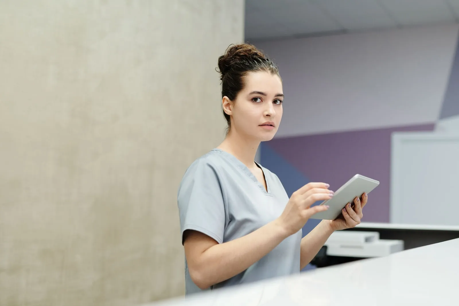 A health worker in scrubs holding a tablet at a clinic reception counter