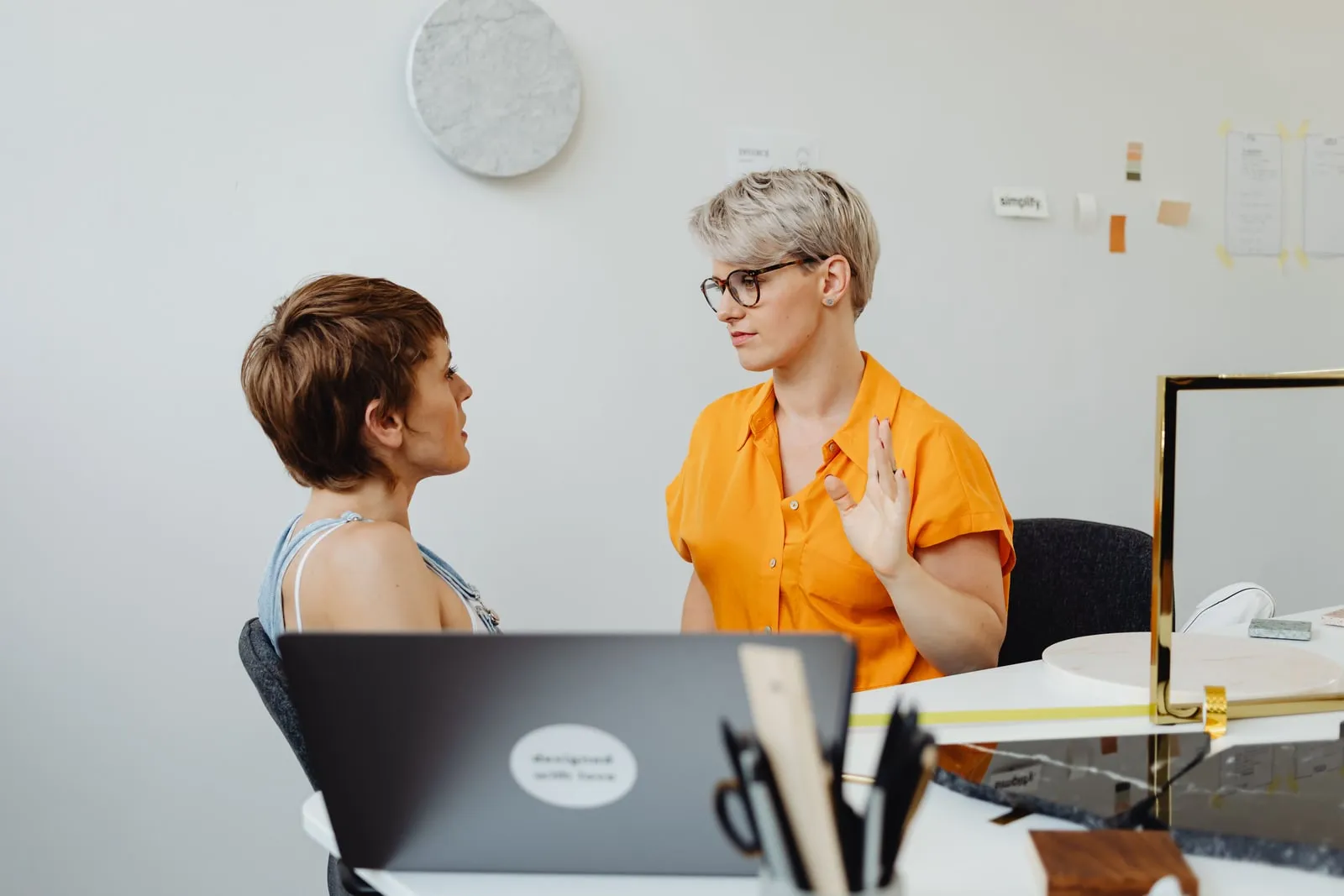 Two colleagues talking at a desk with an open laptop in a bright office