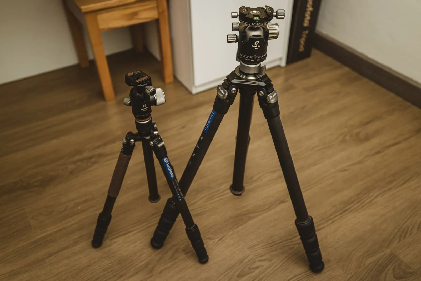 Two camera tripods standing on a wooden floor indoors