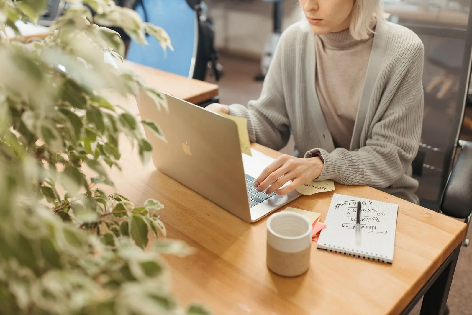 A person typing at a laptop on a wooden desk with notes and a mug beside it