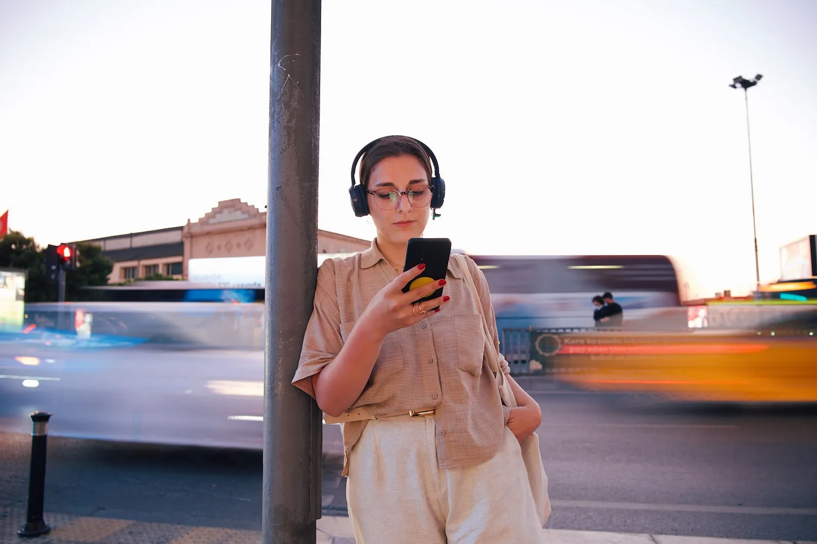 A person in headphones looking at a phone beside a busy road at dusk