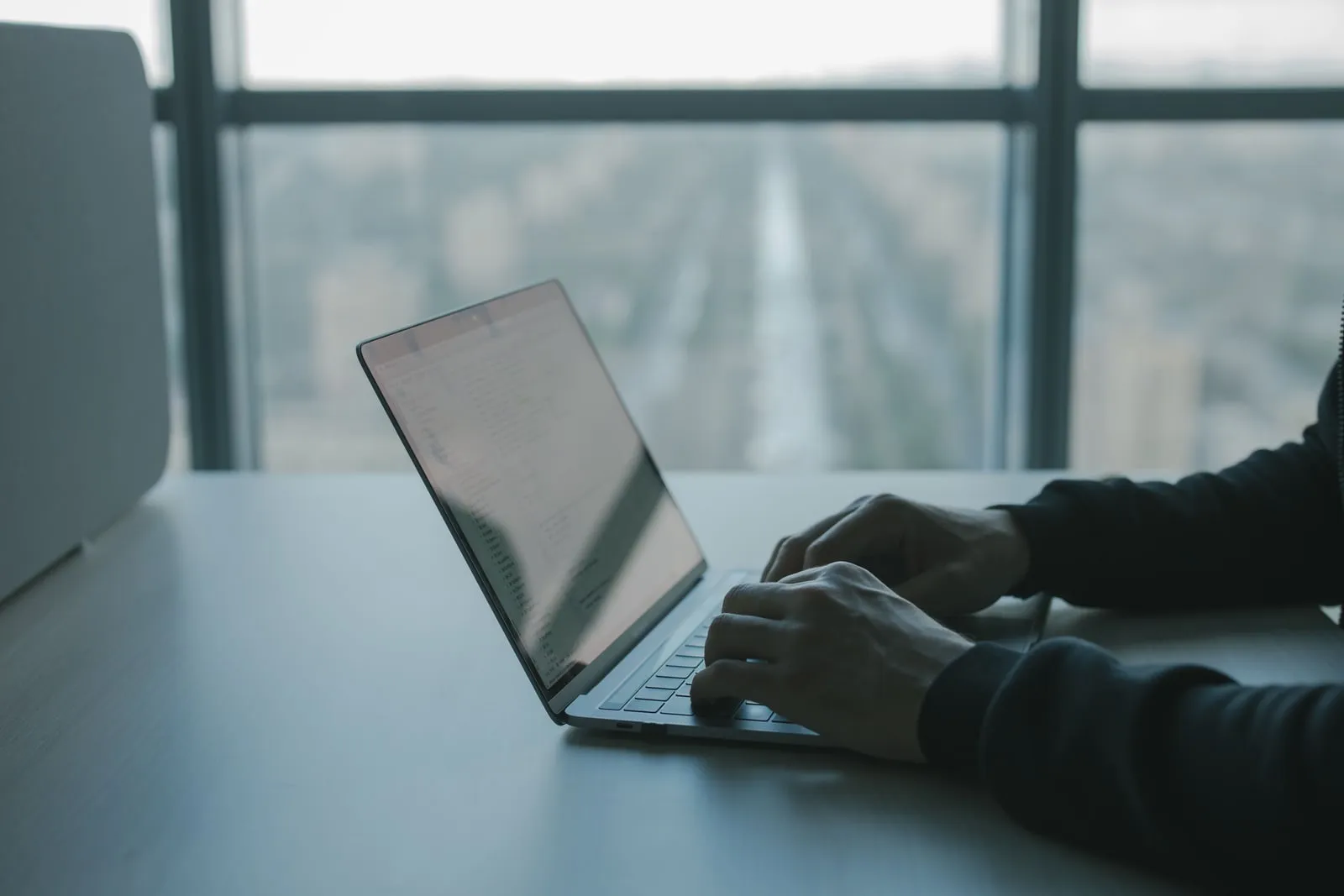 A laptop on a desk by a window, with two hands typing on the keyboard