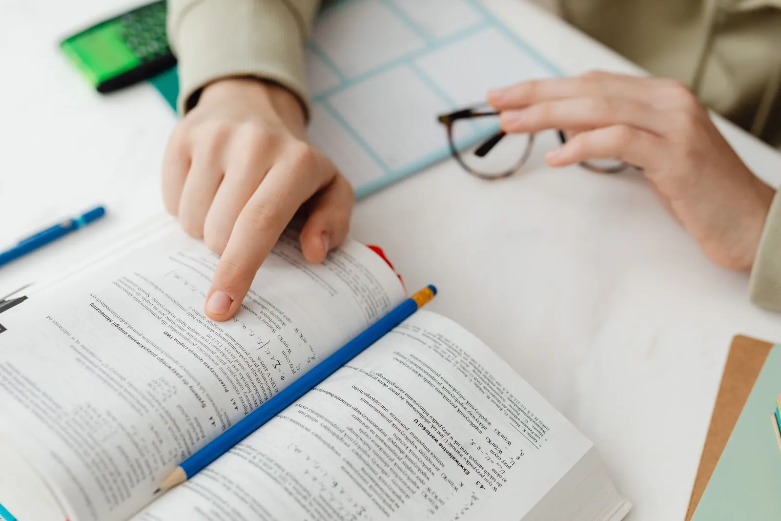 A person pointing at a page of an open textbook while holding a pair of glasses