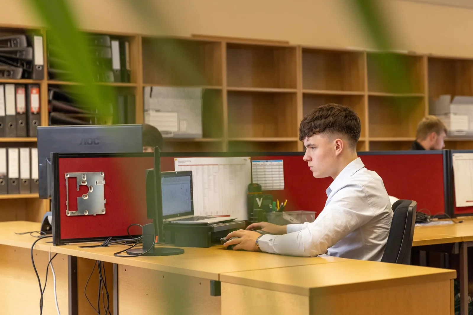 An office worker at a desk with two monitors in a shared administrative office