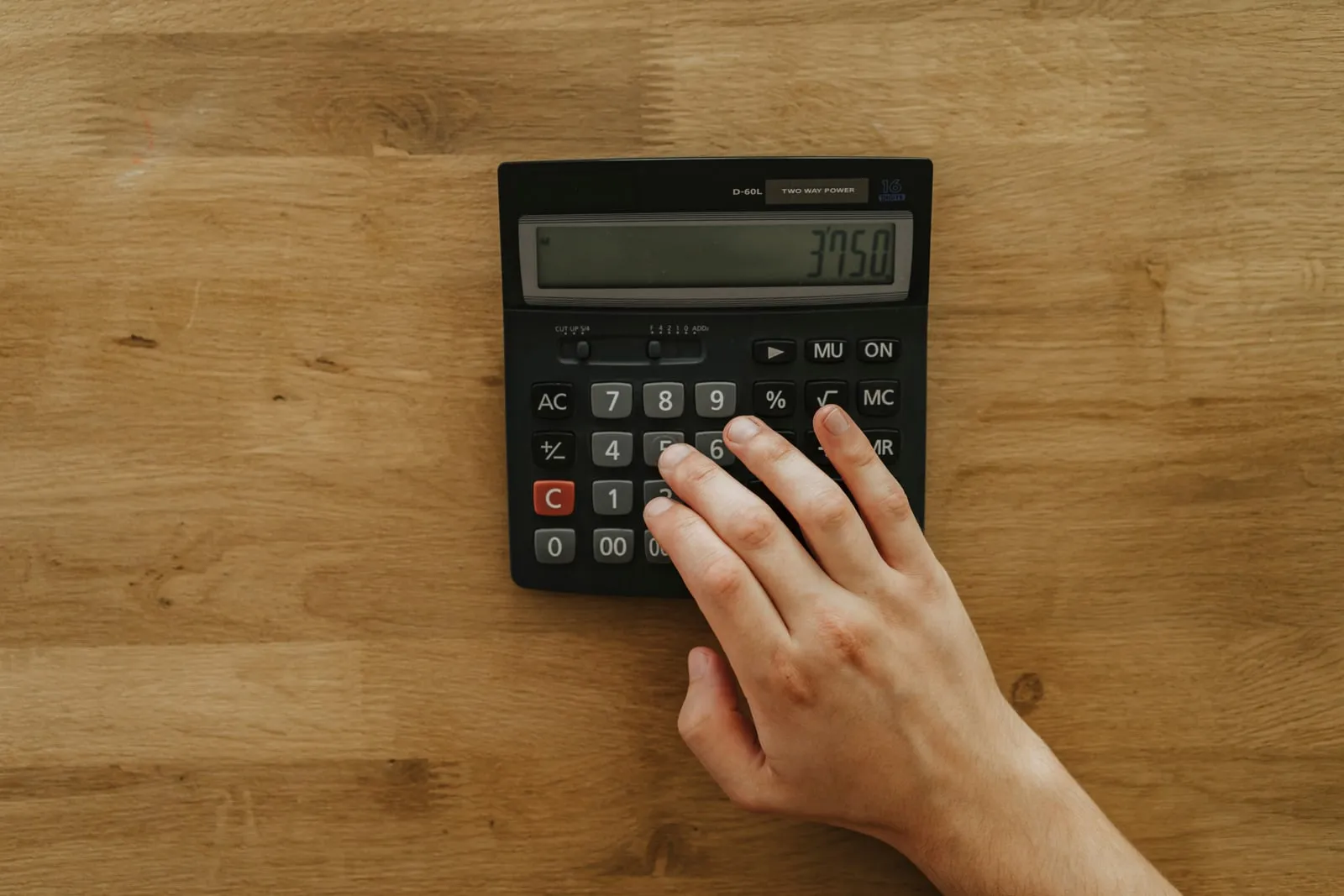 A hand pressing the keys of a desk calculator on a wooden table