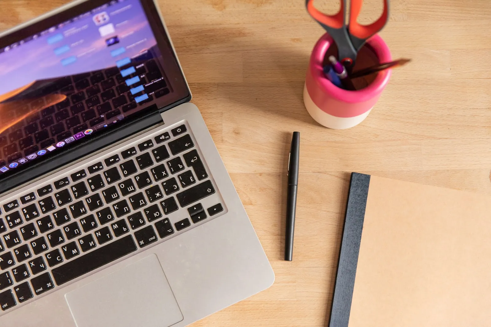 A closed notebook and a pen on a wooden desk beside an open laptop