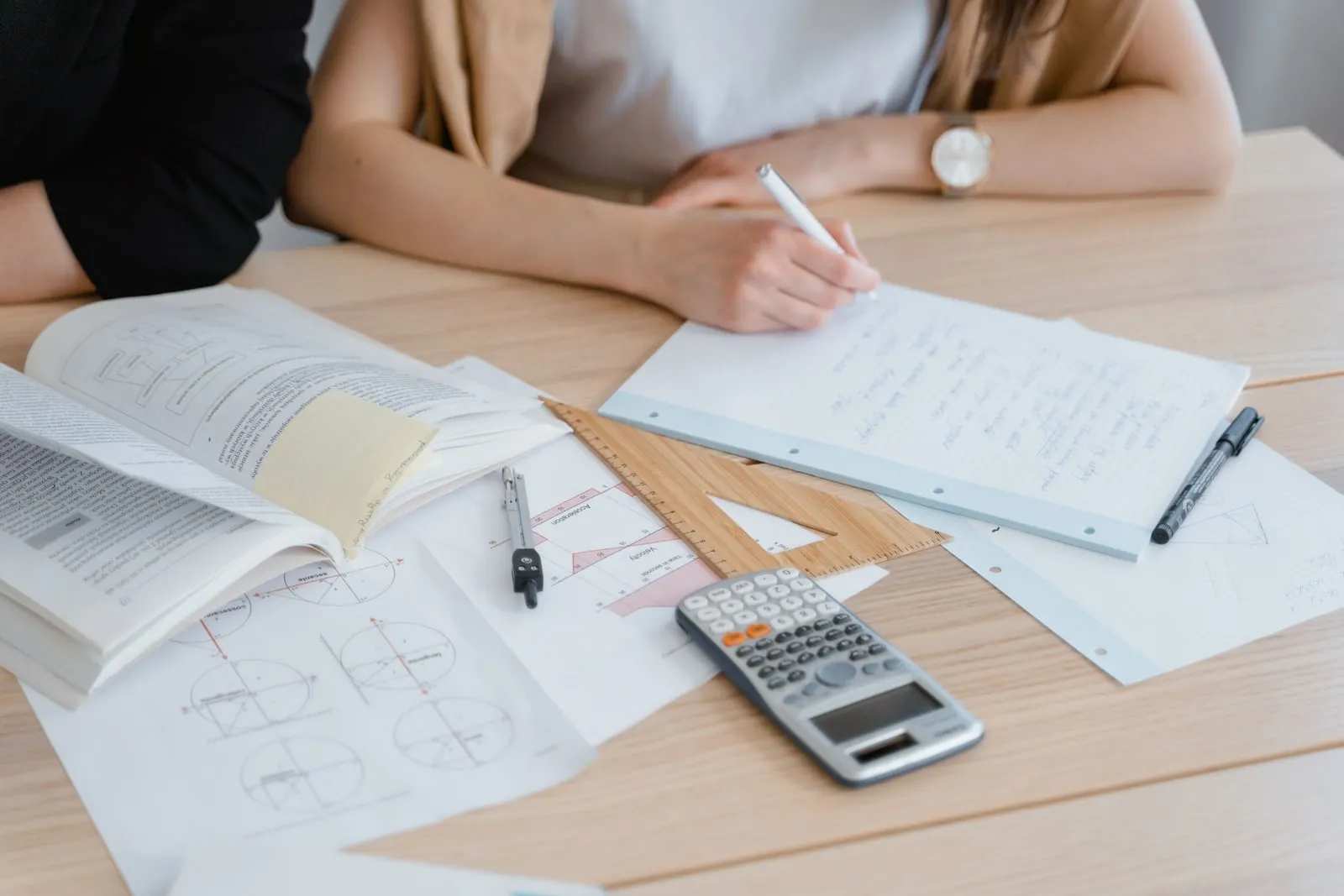 A desk with a maths textbook, compass, ruler, calculator and handwritten working