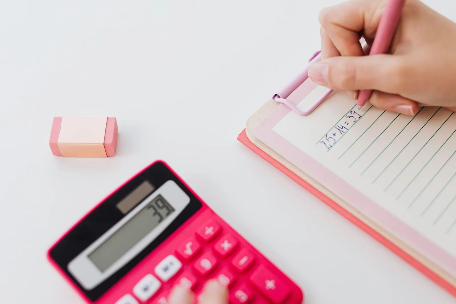 A hand writing a sum in a notebook beside a calculator